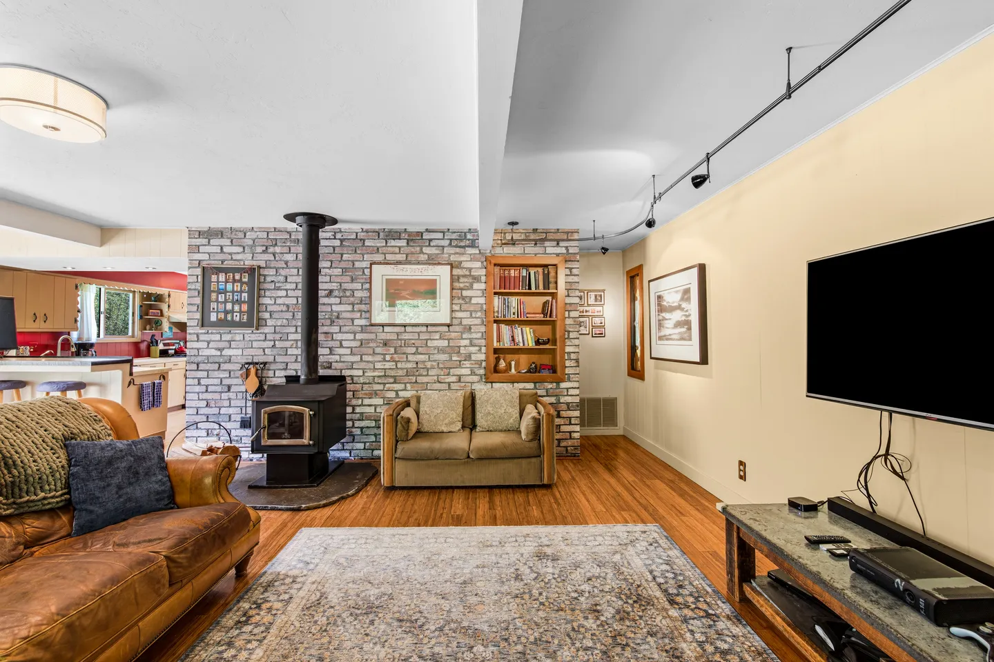 Living room with a wood-burning stove against a brick accent wall, hardwood floors, seating, and a mounted flat-screen television.