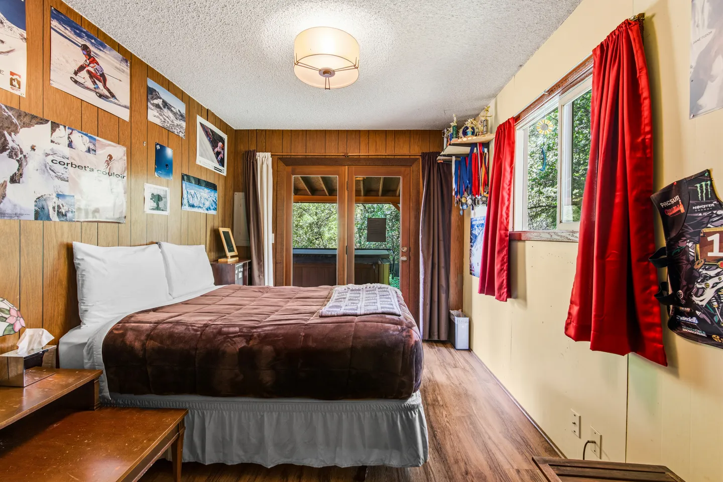 Bedroom with wood-paneled walls, ski posters, a bed with a brown blanket, and glass doors leading outside.