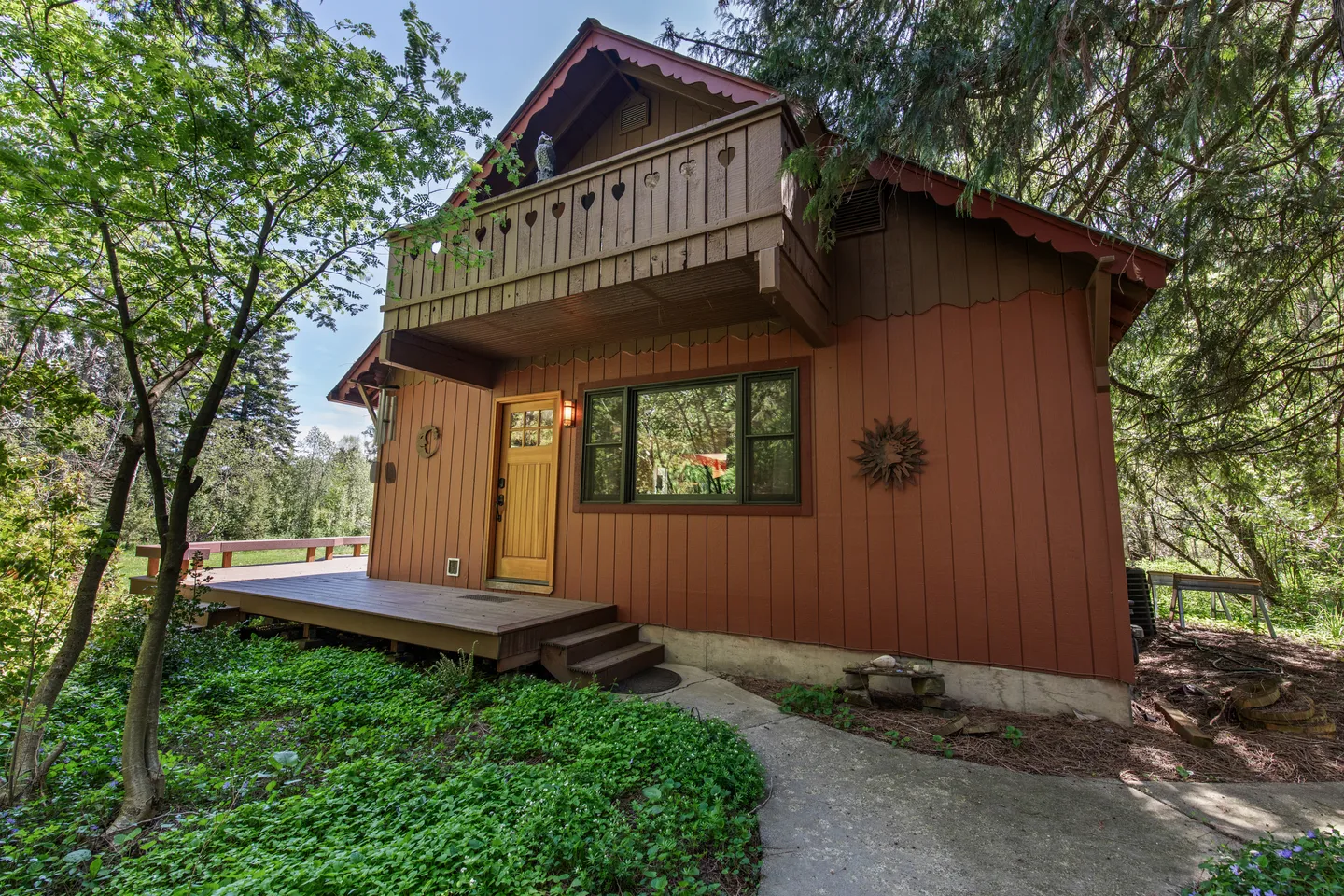 Exterior of a two-story chalet-style cabin with red-brown siding, a front porch, and an upper wooden balcony with cutout details.