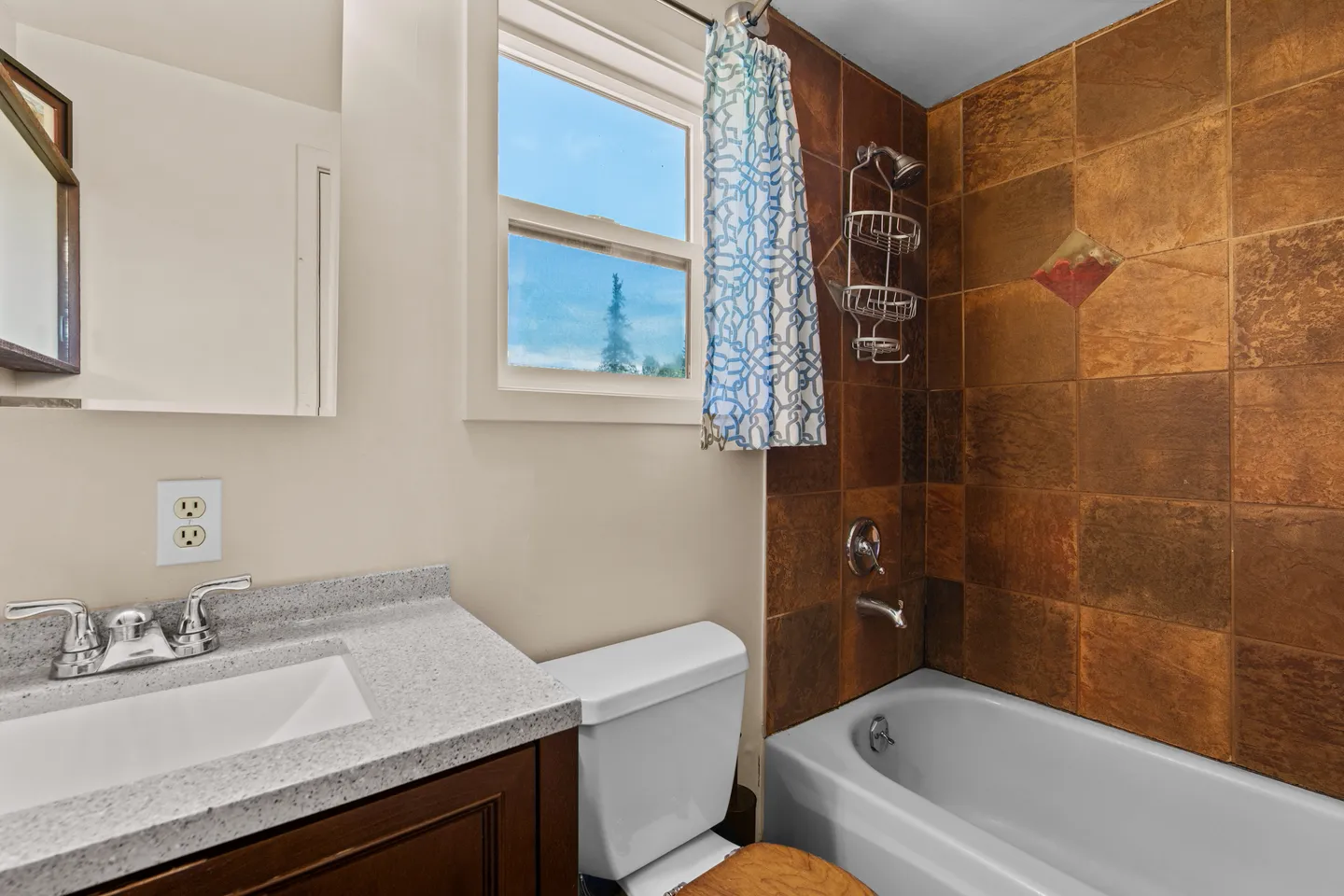 Bathroom with vanity, toilet, and bathtub with warm brown tile surround next to a window.