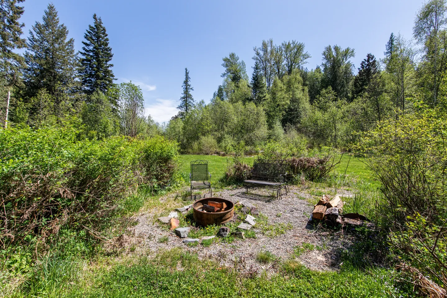 Outdoor gravel fire pit area with a metal ring, firewood, a bench, and a chair surrounded by greenery and trees.