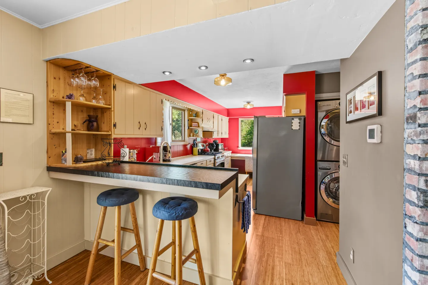 Kitchen view looking past a raised breakfast bar with two cushioned stools toward wooden cabinetry, red walls, and stacked laundry appliances.