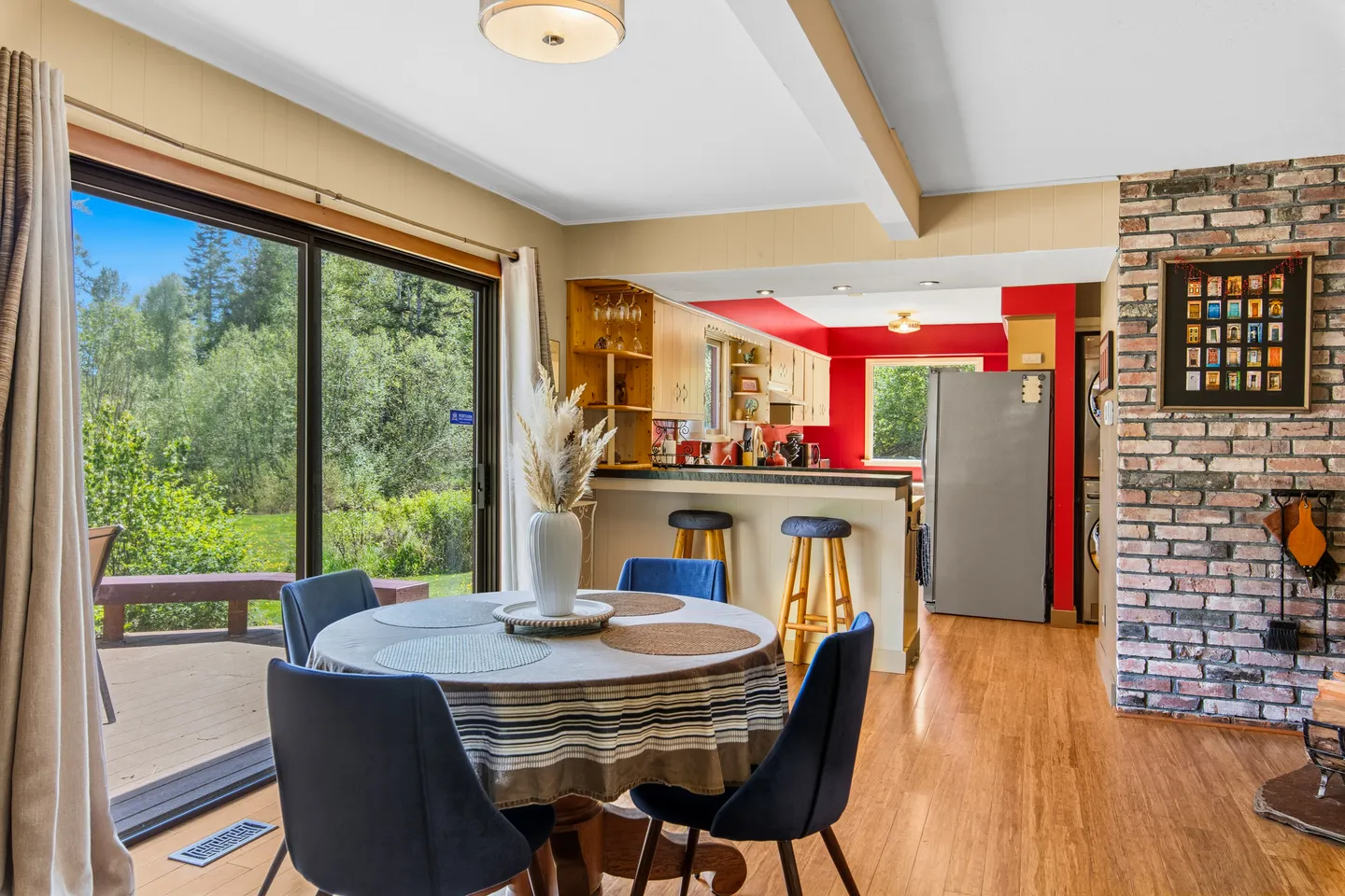 Dining area with a round table and blue chairs beside sliding glass doors leading to an outdoor deck, adjacent to an open kitchen with a breakfast bar.