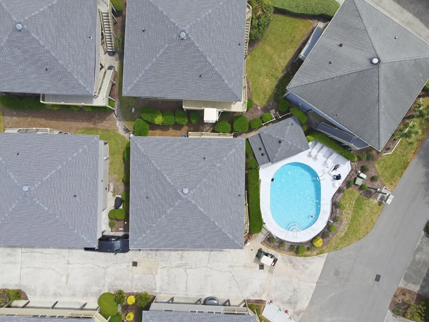 Bird's-eye aerial view of residential roofs, paved driveways, landscaping, and a fenced swimming pool with sun loungers.