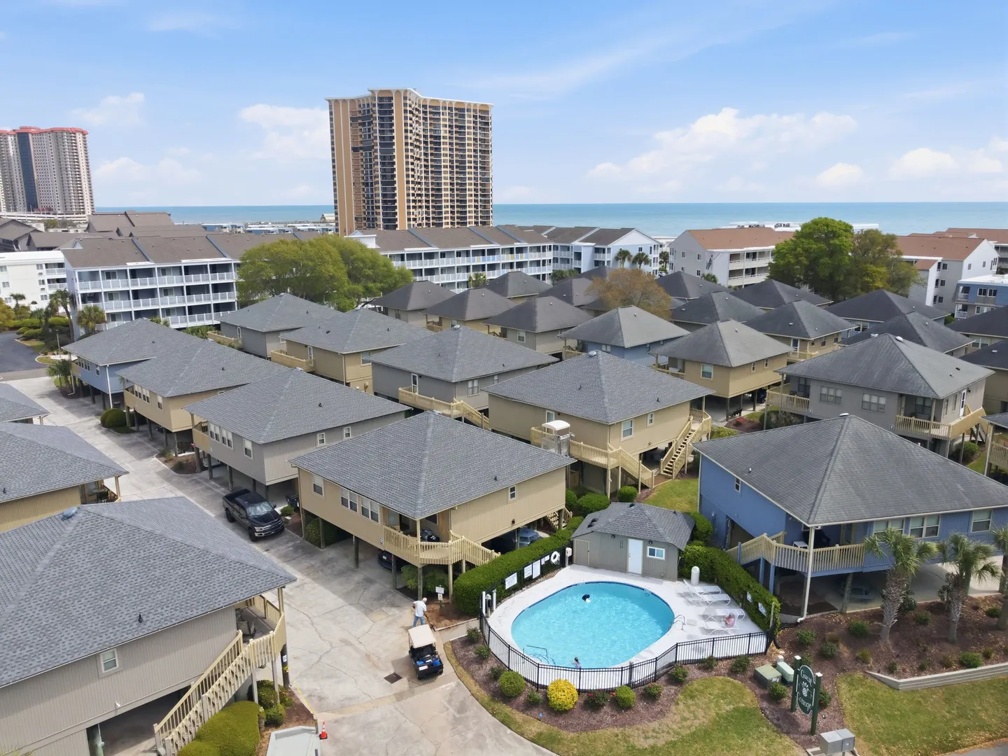 Aerial view of a coastal neighborhood with stilt homes, a community swimming pool, and high-rise buildings near the ocean.