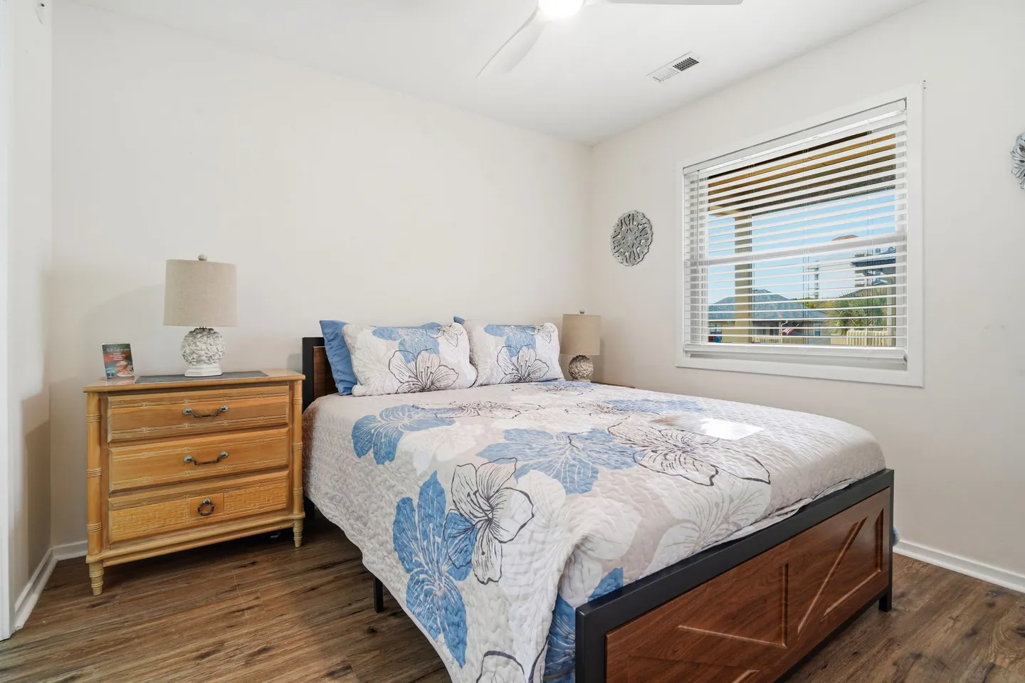Bedroom with a wooden bed frame, blue floral quilt, light wood dresser, and a window with blinds.