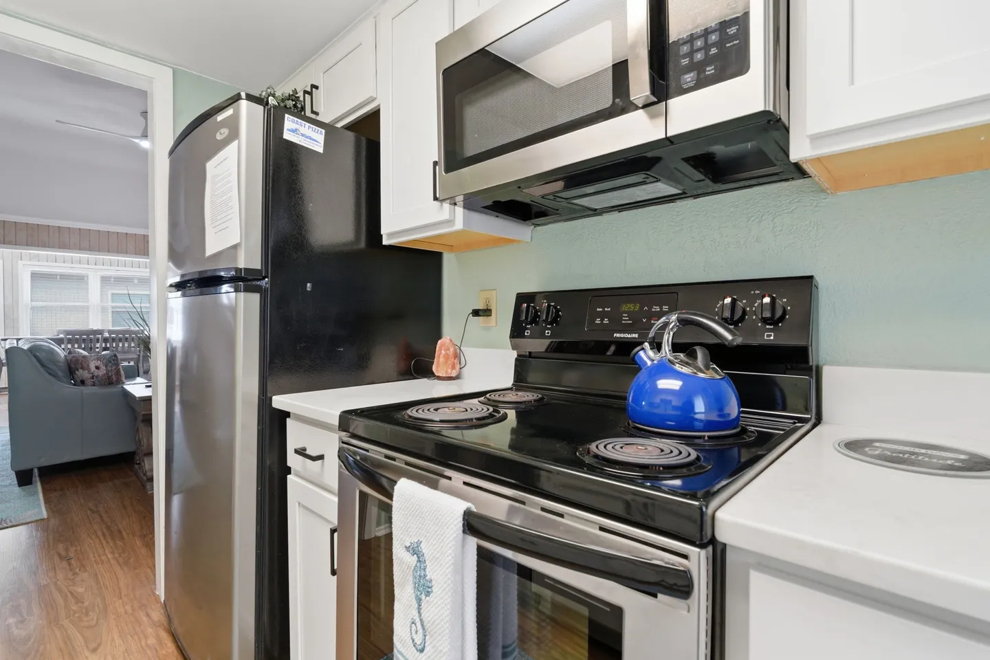 Close-up view of a kitchen featuring a stainless steel stove with a blue kettle, an over-the-range microwave, and a refrigerator next to white cabinets.