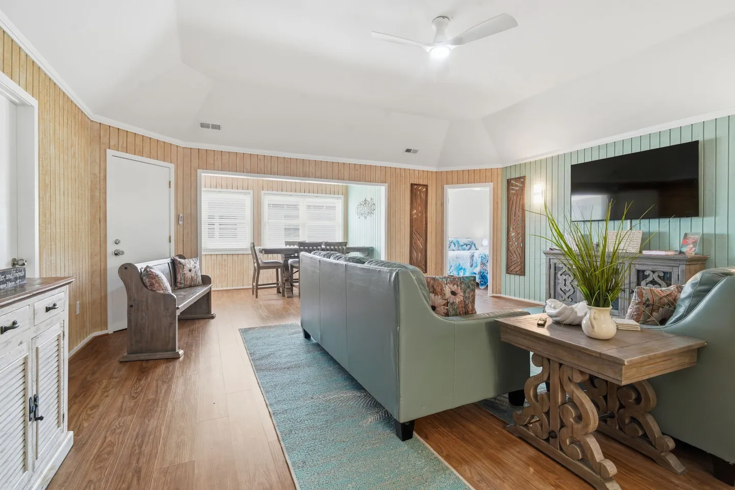Living room featuring paneled walls, a high angled ceiling, blue sofas, a wall-mounted TV, and an open entryway to an adjacent dining area.