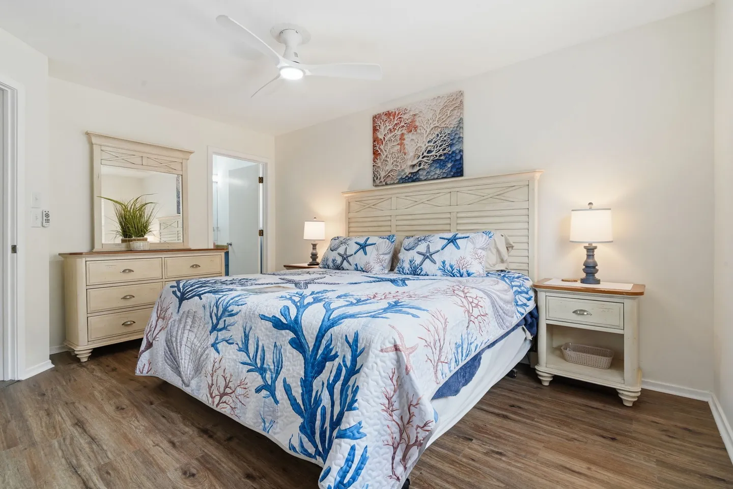 Bedroom with coastal-themed coral bedding, a white headboard, matching nightstand and dresser, and wood-look flooring.