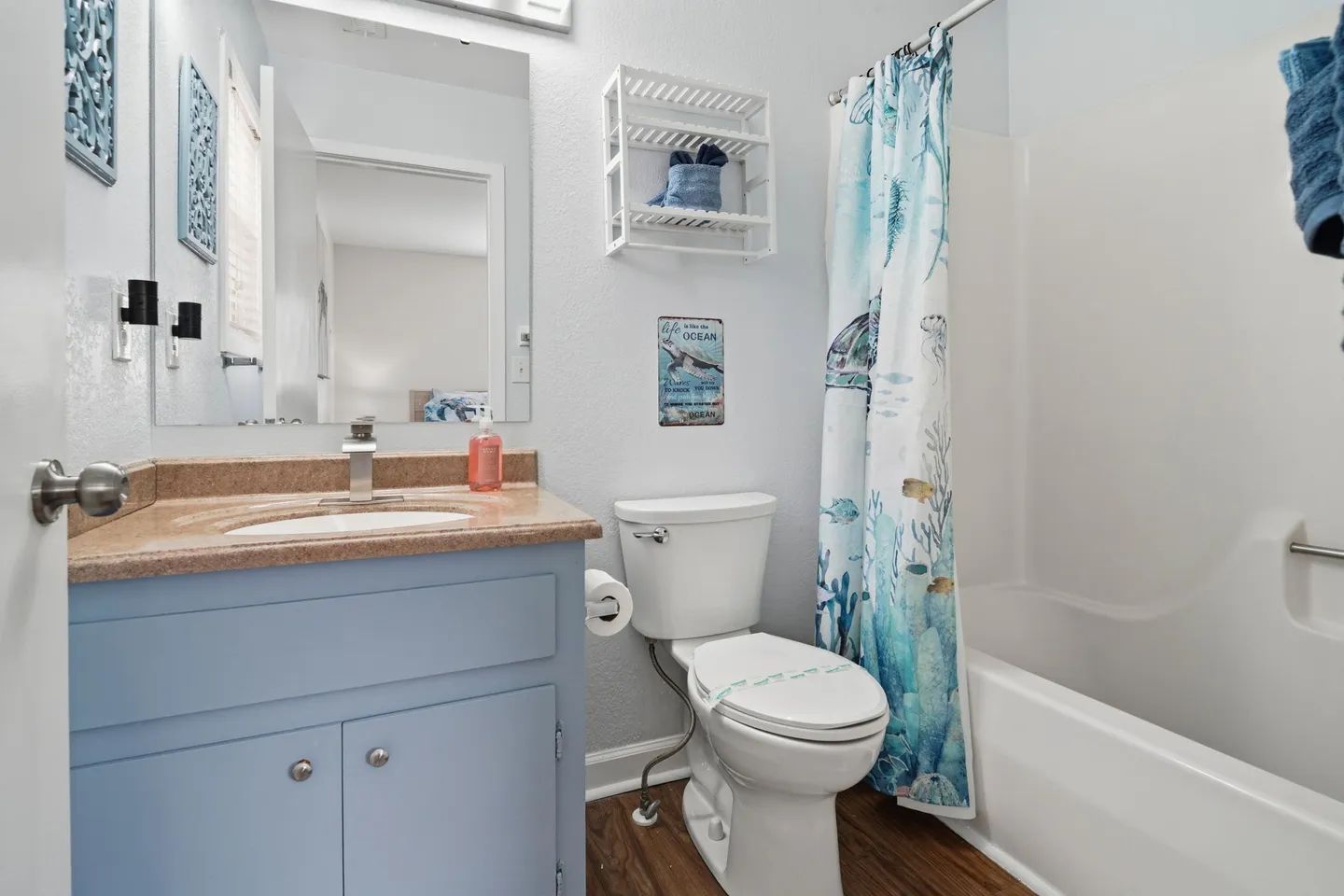 Bathroom with a blue vanity, toilet, bathtub shower combo with an ocean-themed curtain, and small white wall shelf.