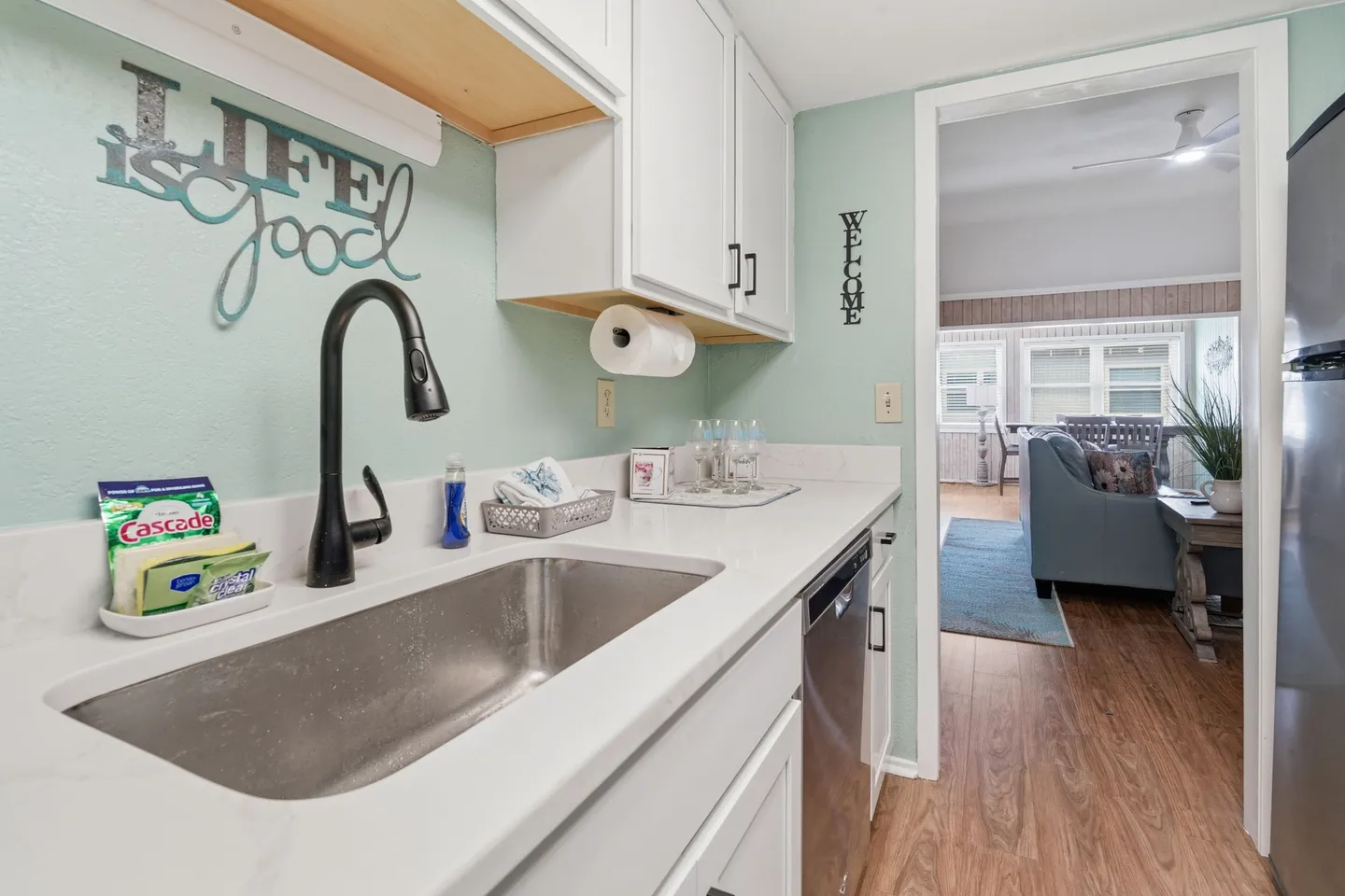 Close-up of a kitchen sink with a gooseneck faucet, light countertops, and white cabinets, looking through a doorway into the living area.