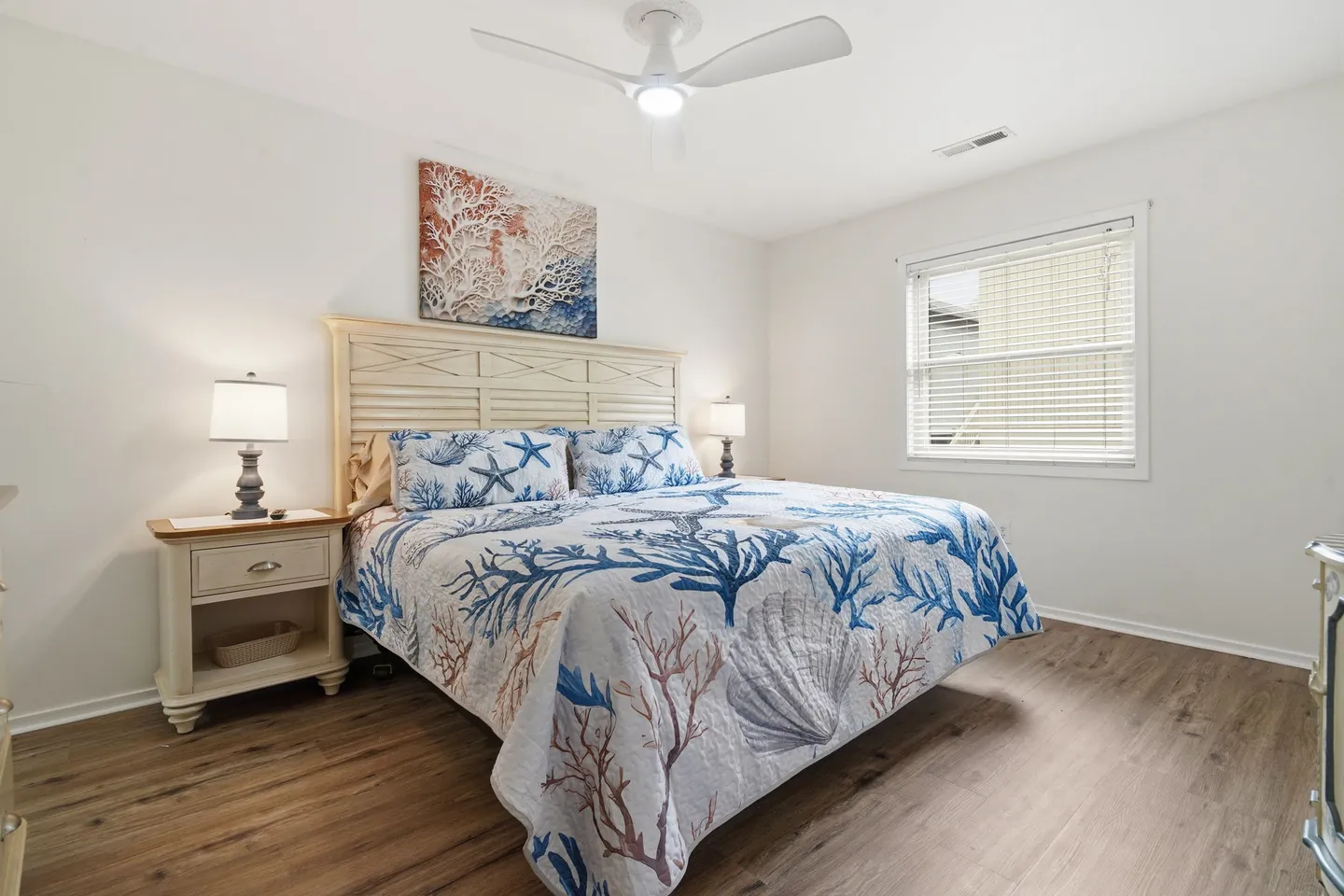 Bedroom with coastal-themed bedding, a white headboard, nightstands with lamps, wood-look flooring, and a ceiling fan.