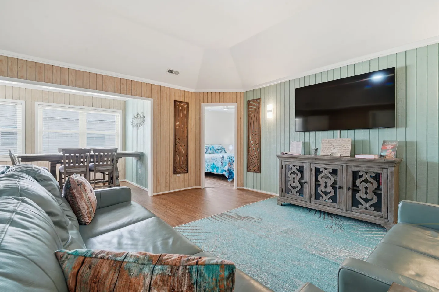 Living room with blue leather couches, an area rug, a TV mounted on a green paneled accent wall above a wooden console, and open doorways to a dining area and bedroom.