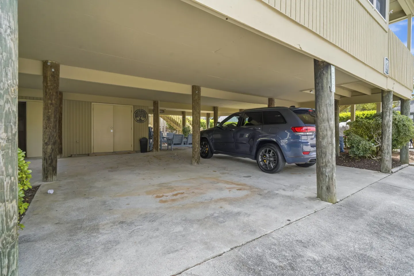 Covered concrete carport beneath an elevated stilt home with wooden support pilings and a parked SUV.
