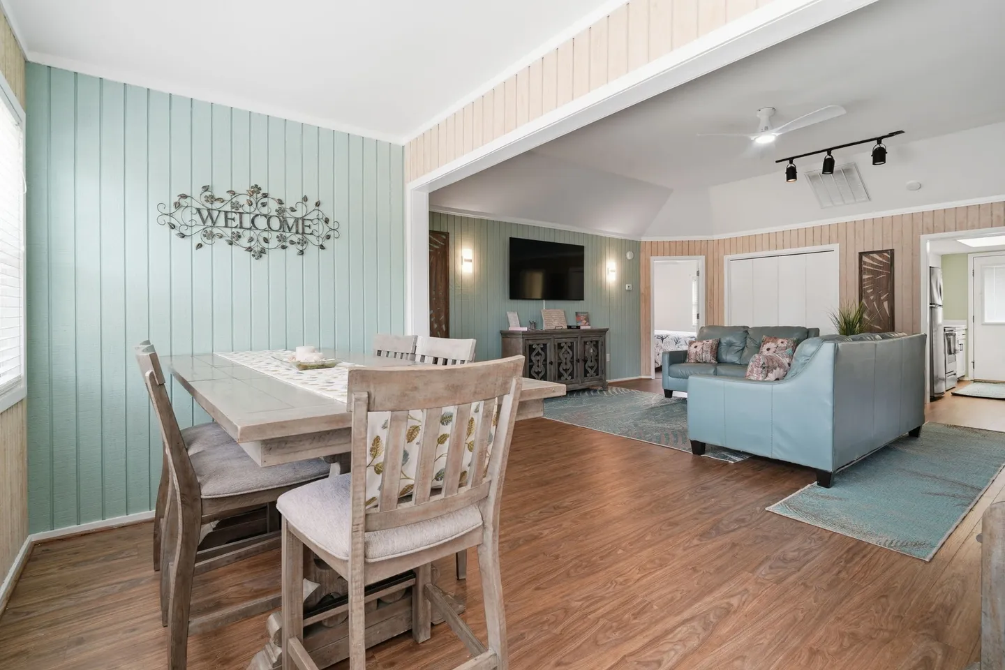 Open-concept dining and living area with wood-look flooring, a dining table with chairs, and a living room with a sectional sofa and mounted TV.