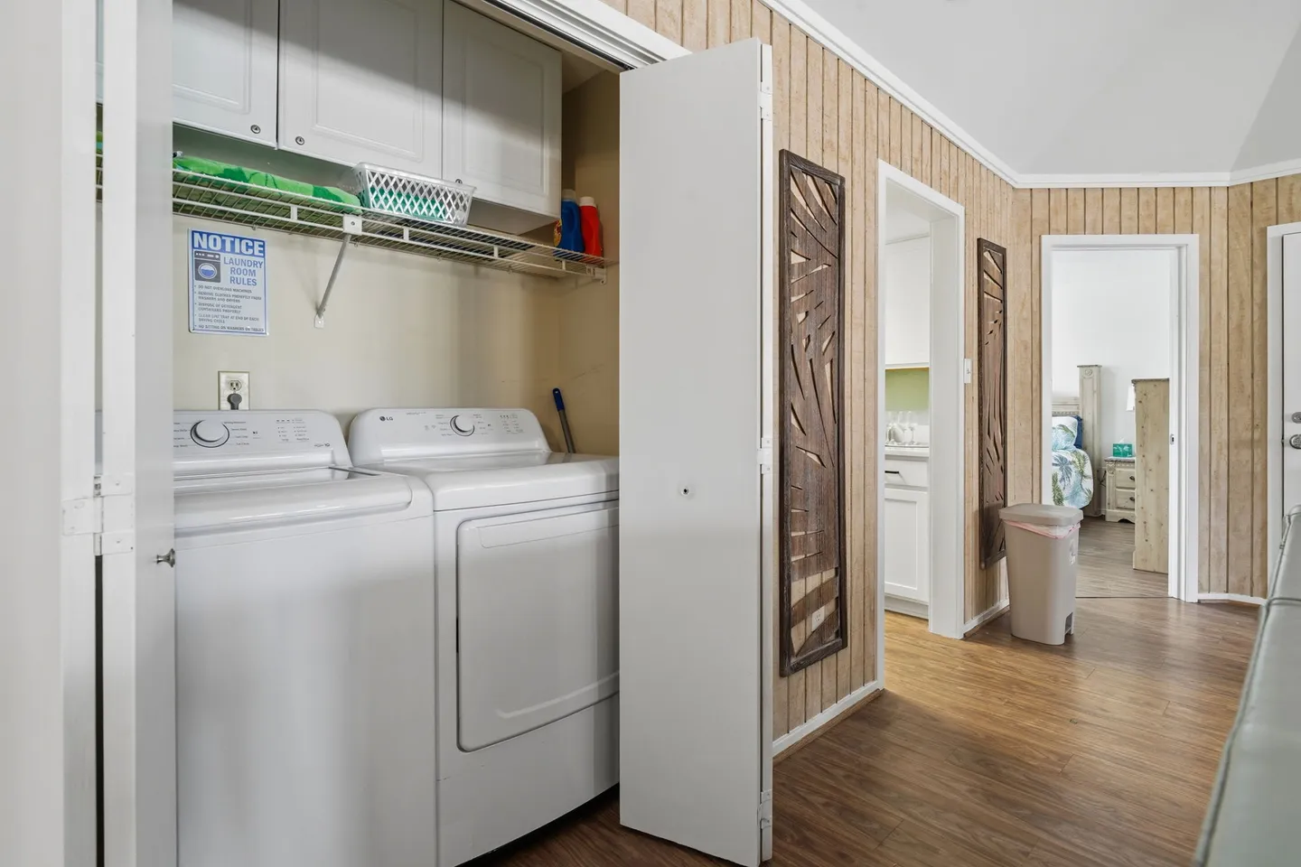 Open closet revealing a white washer and dryer with upper cabinets and wire shelving, located in a hallway with wood-look paneled walls.