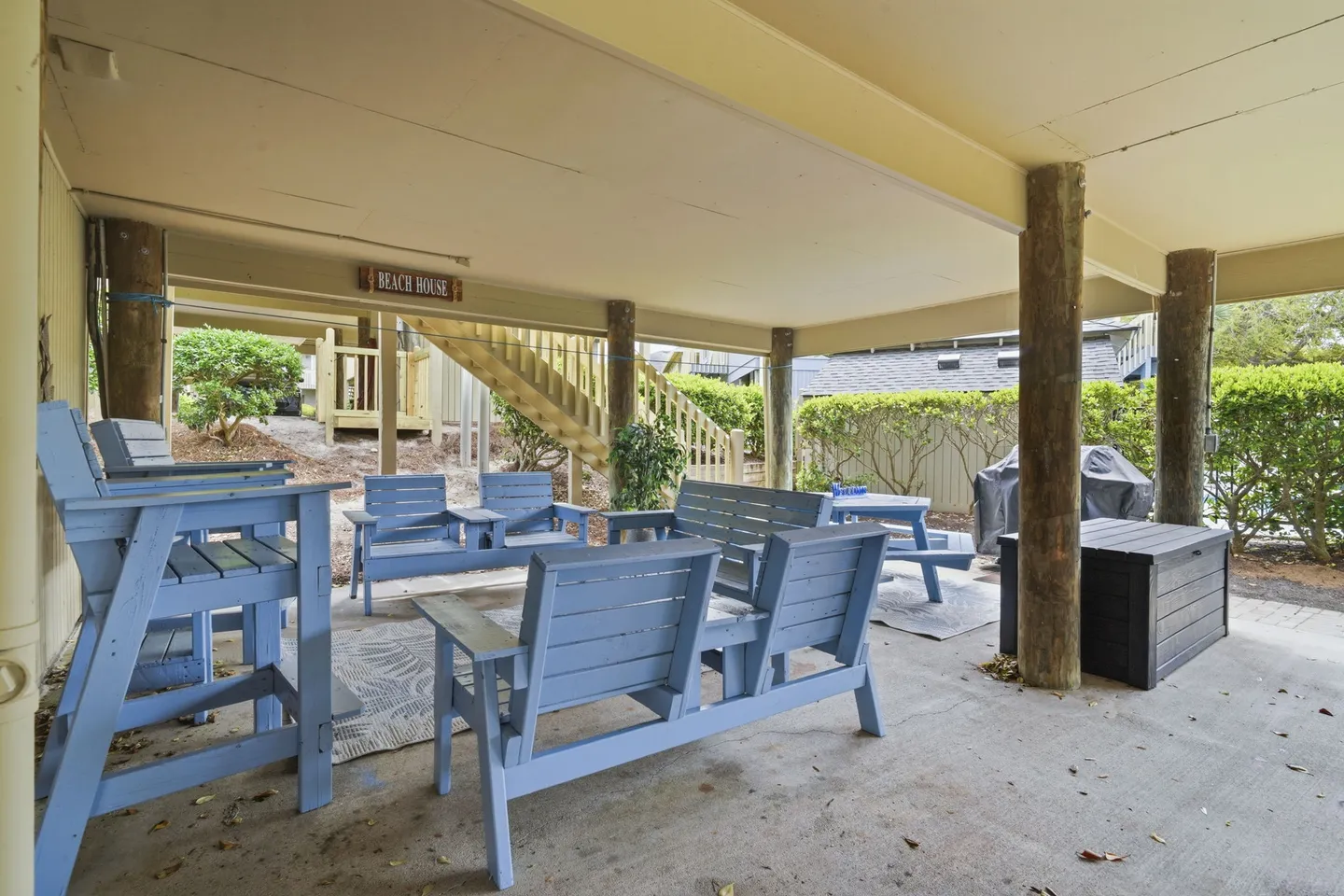 Covered ground-level patio beneath a stilt home featuring light blue wooden seating, a picnic table, and outdoor stairs.