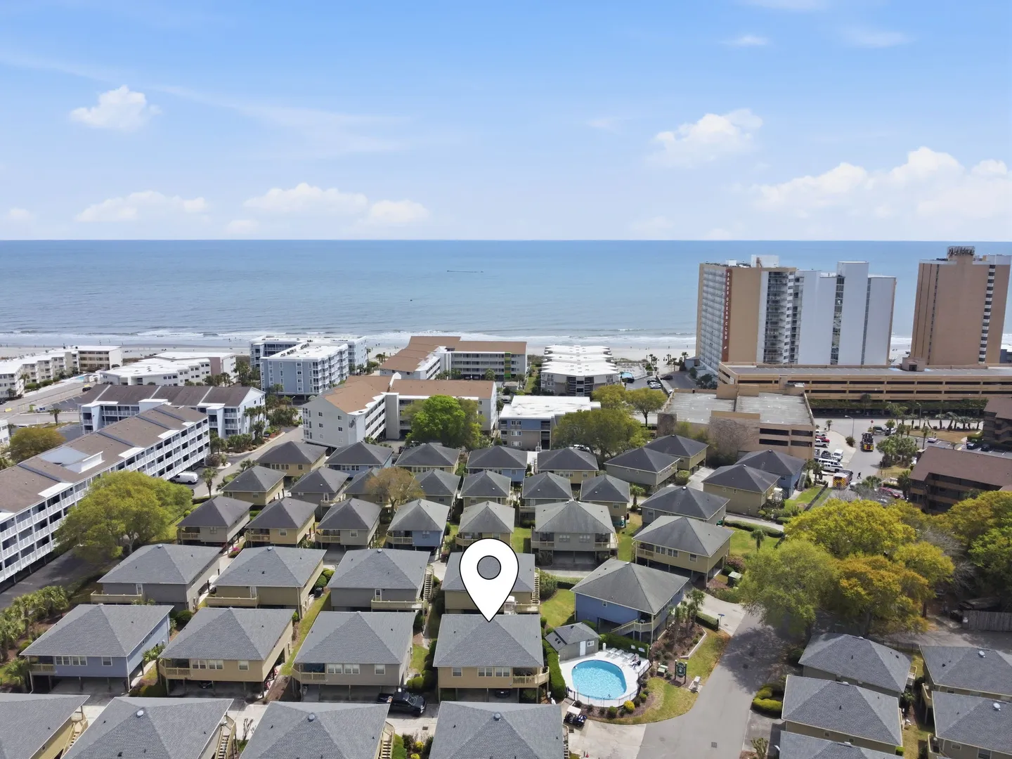 Aerial view of a coastal neighborhood with pitched-roof houses, an outdoor pool, and the ocean in the background under a blue sky.