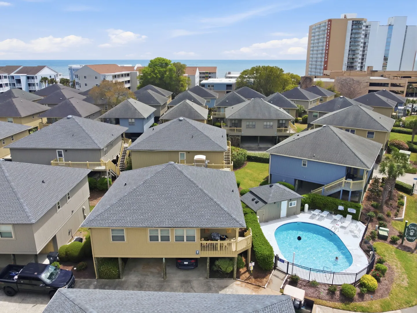 Aerial view of a coastal cottage community with raised stilt homes, a community swimming pool, and the ocean visible in the background.