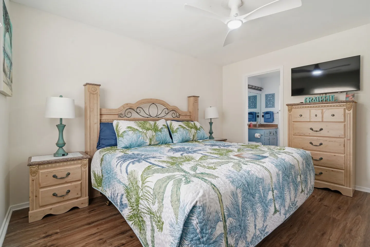 Bedroom with a wooden bed dressed in palm-leaf bedding, flanked by nightstands and facing a dresser with a wall-mounted TV.