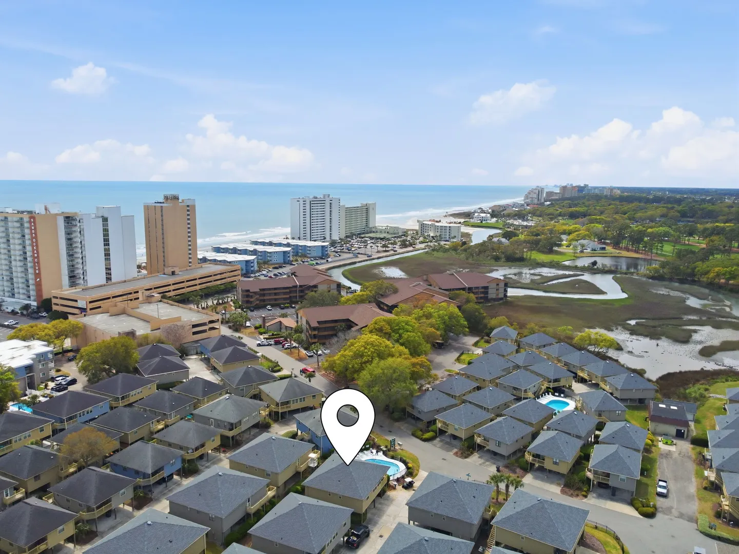 Aerial view of a coastal neighborhood with townhomes, shared swimming pools, and the ocean in the background with a location pin marker.