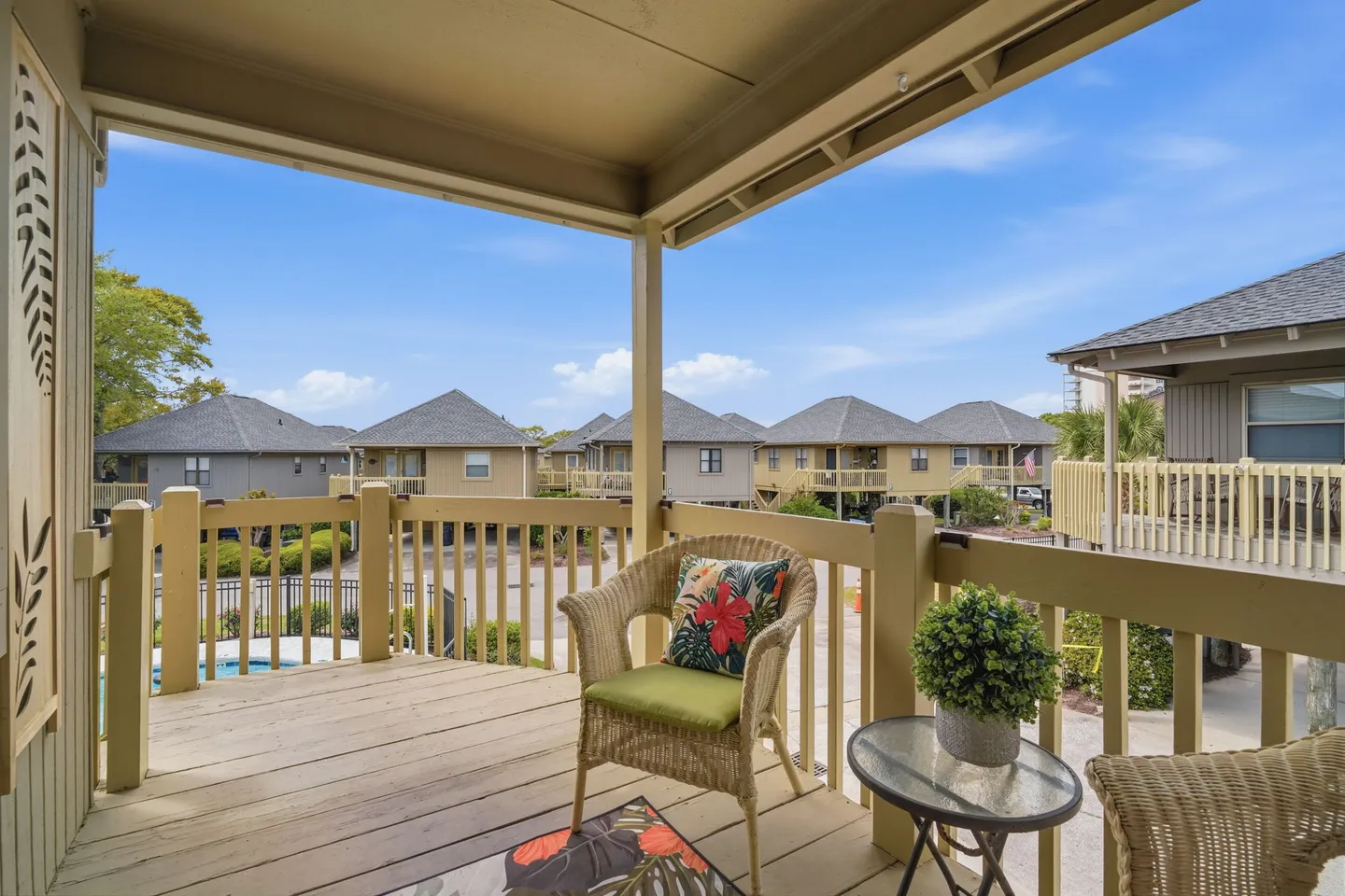 Covered outdoor balcony with wicker chairs, a small table, wooden railings, and views of neighboring houses and blue sky.