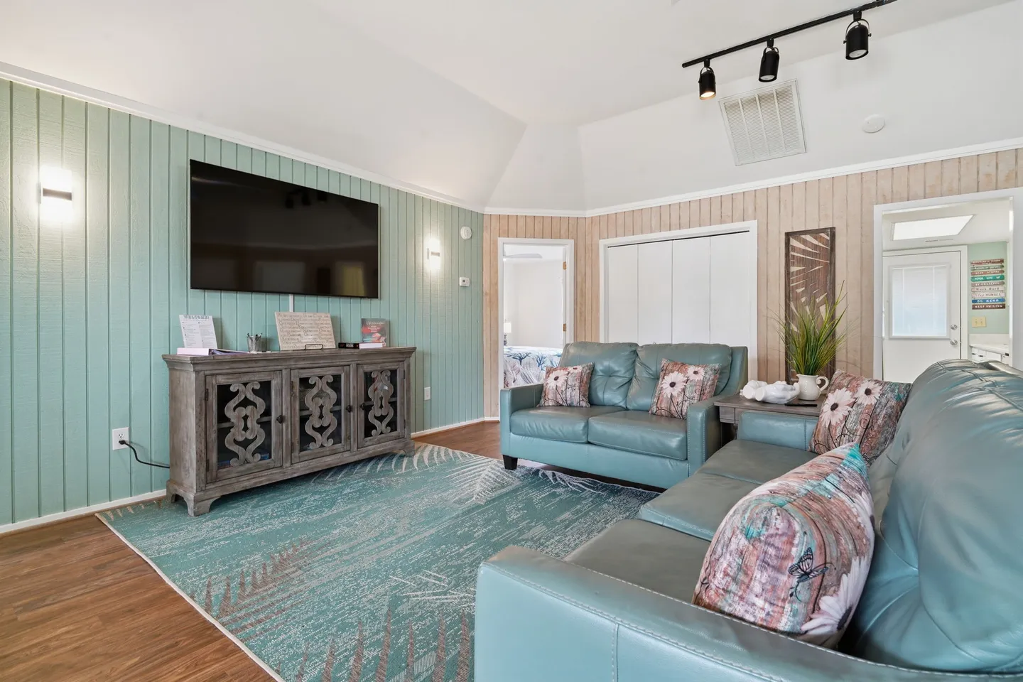 Living room with light blue-green paneled accent wall, wall-mounted television over a carved console, and teal leather seating over a patterned rug.