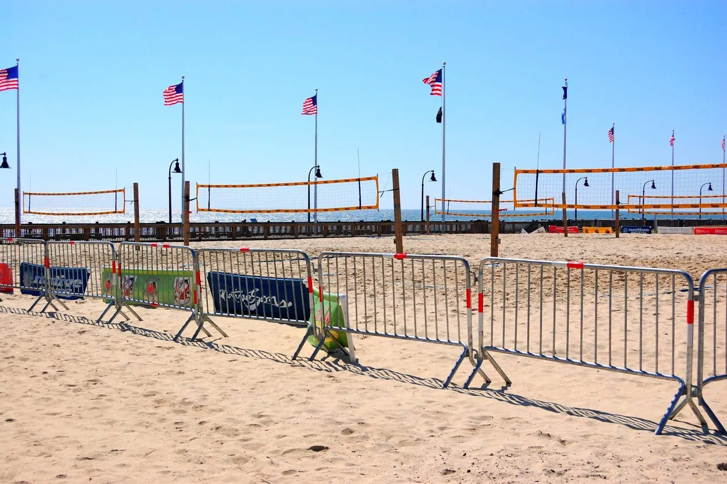 Beach volleyball courts set up on the sand with metal barricades in the foreground and American flags flying under a blue sky.