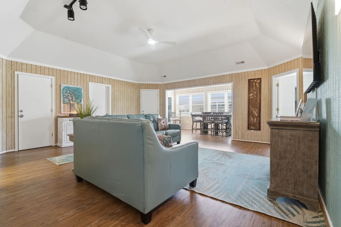 Living room with light blue sofas, wood-planked walls, vaulted white ceiling, and an adjoining enclosed dining area.