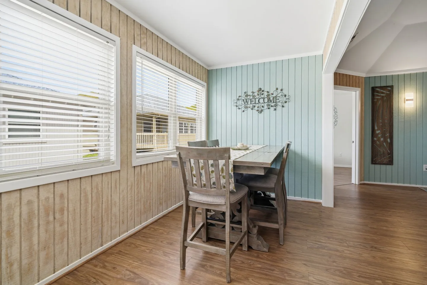 Dining nook with a high-top table and chairs next to panel-lined walls and two horizontal-slat shuttered windows.
