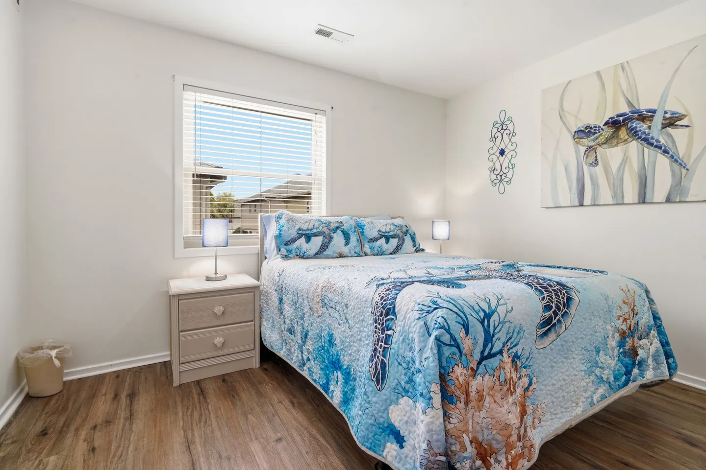 Bedroom with a sea turtle themed quilt, wood-style floors, a window with blinds, and a nightstand with a lamp.