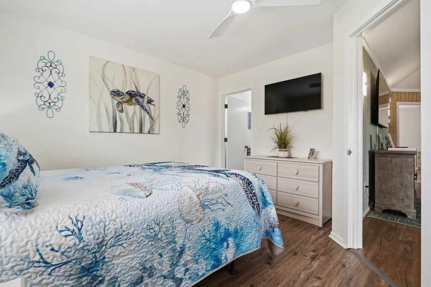 Bedroom with sea turtle and jellyfish themed quilt, wall-mounted TV, white dresser, and wood-look flooring.