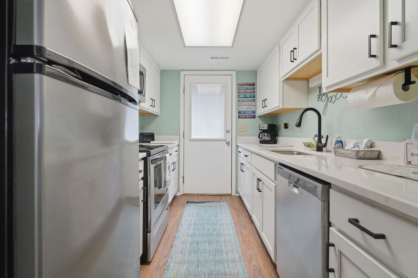 A galley kitchen with white cabinets, stainless steel appliances, light countertops, and an exterior door at the far end.