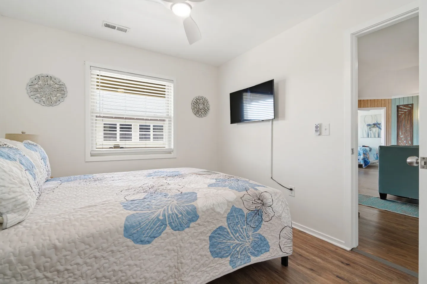 Bedroom with a floral patterned bedspread, a wall-mounted flat-screen TV, a window with blinds, and wood-look flooring.