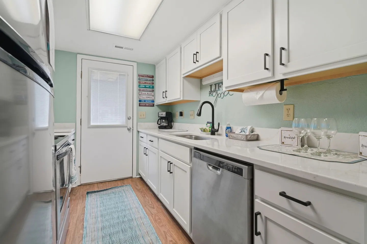 Galley kitchen featuring white cabinets, light countertops, stainless steel dishwasher, and an exterior door at the end.