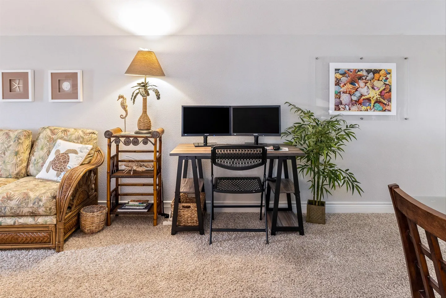 A dedicated workstation in a carpeted room featuring a wooden trestle desk, two monitors, an office chair, and coastal decor.