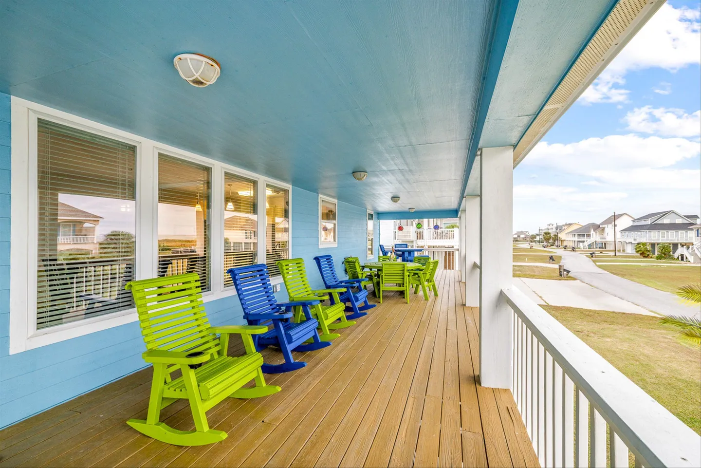 A long covered porch with wooden deck flooring, blue ceiling, and bright green and blue rocking chairs overlooking the neighborhood.