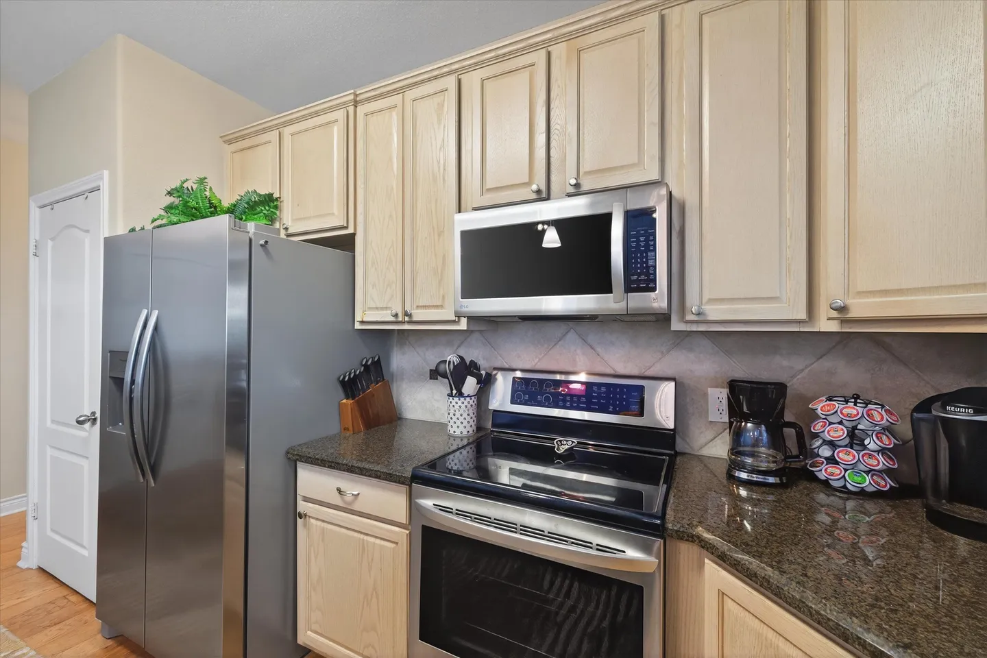 Kitchen counter area with light wood cabinets, stainless steel appliances including a refrigerator, range, microwave, and small coffee makers on the counter.