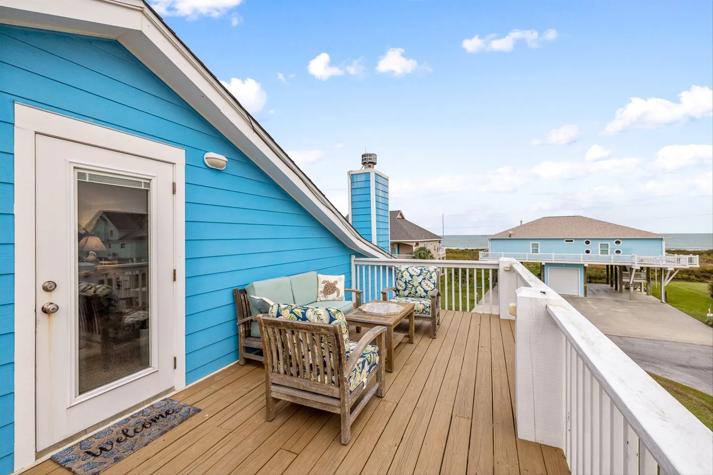Elevated wooden deck with outdoor seating against bright blue siding, offering distant ocean views under a blue sky.