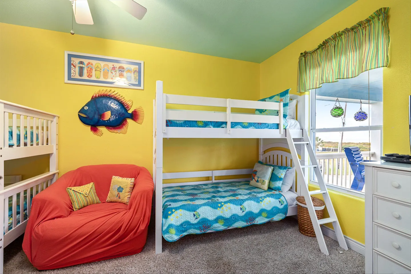 Bedroom with yellow walls, a teal ceiling, white bunk beds with fish-themed bedding, a red armchair, and a window.