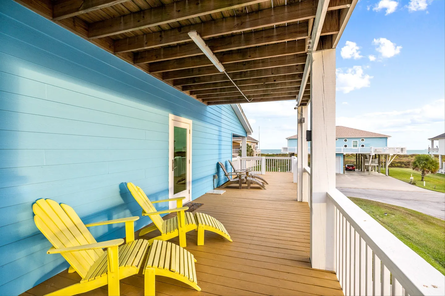 Covered wooden deck with bright yellow lounge chairs against a light blue exterior wall overlooking a neighborhood and distant ocean view.