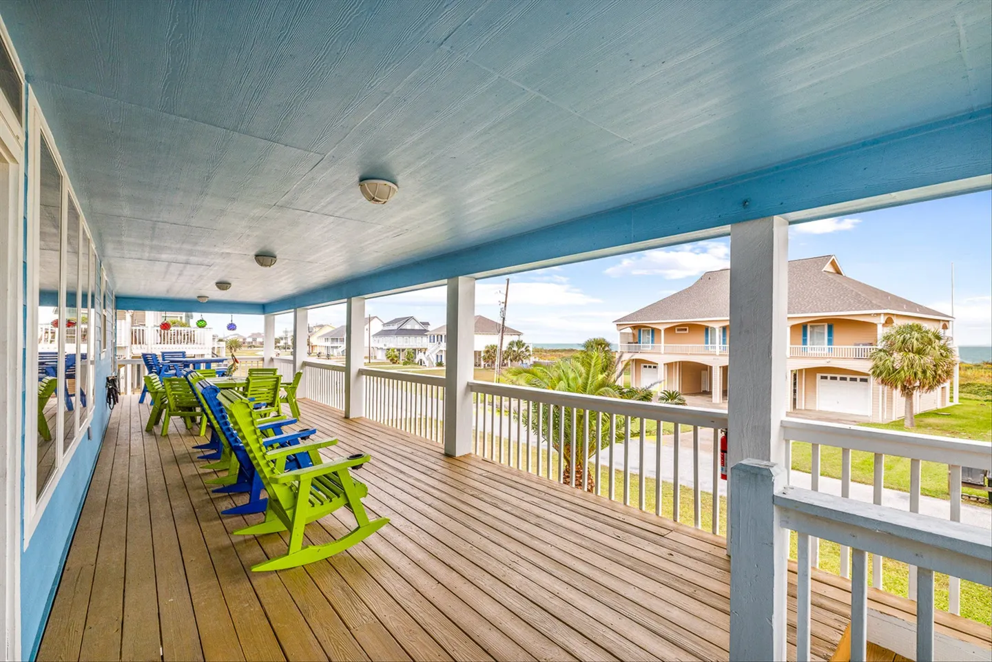 A covered deck with wooden flooring, light blue ceiling, and colorful green and blue rocking chairs overlooking neighboring beach homes.