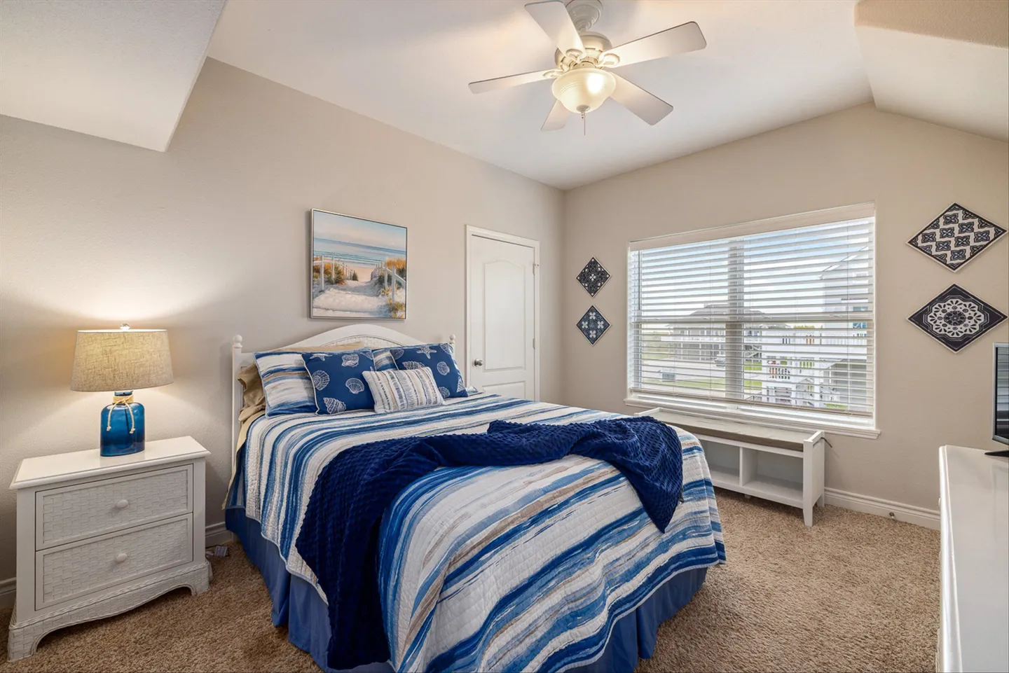 Bedroom with a bed made up in blue and white coastal bedding, a white nightstand, and a window with horizontal blinds.
