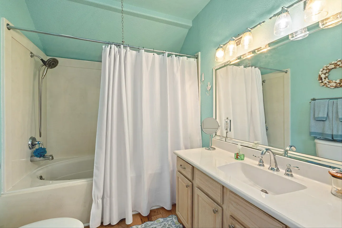 Bathroom with light blue walls, a bathtub-shower combination behind a white curtain, and a vanity with a large mirror and overhead lighting.