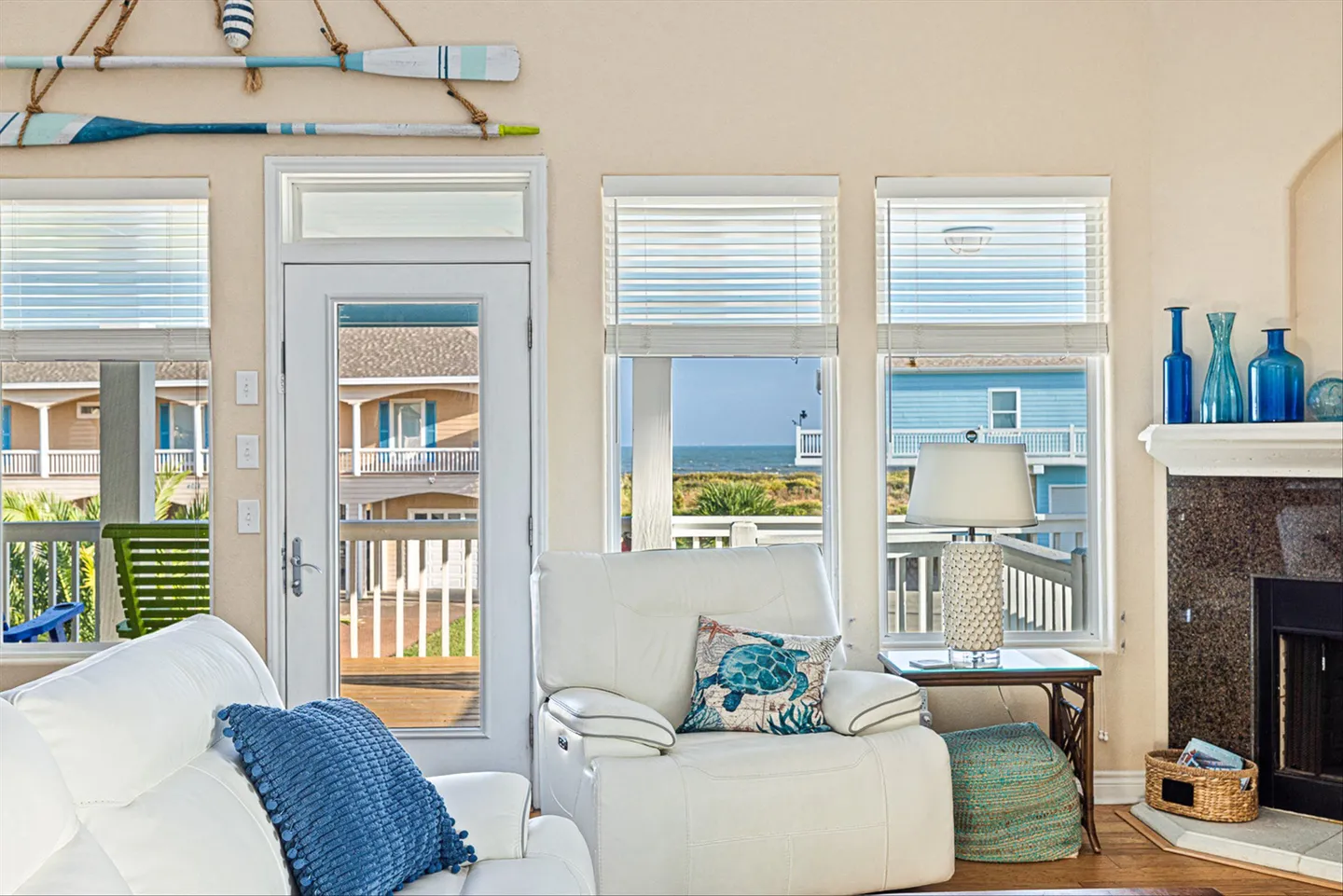Living area with white leather seating, a glass-paneled exterior door, and windows revealing a glimpse of the ocean and adjacent homes.