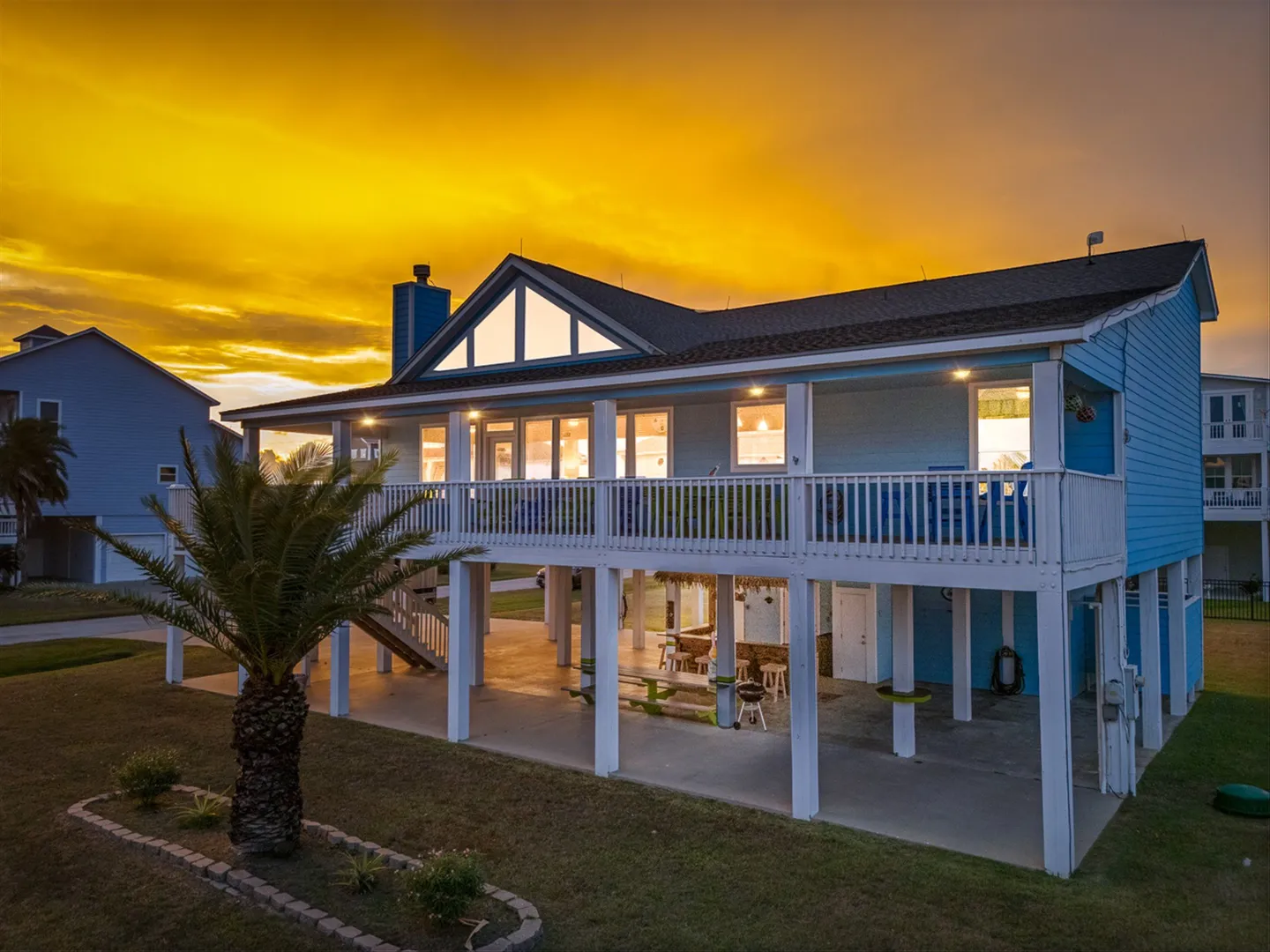 Exterior dusk view of a stilted light blue beach house with a vibrant yellow-orange sunset sky.