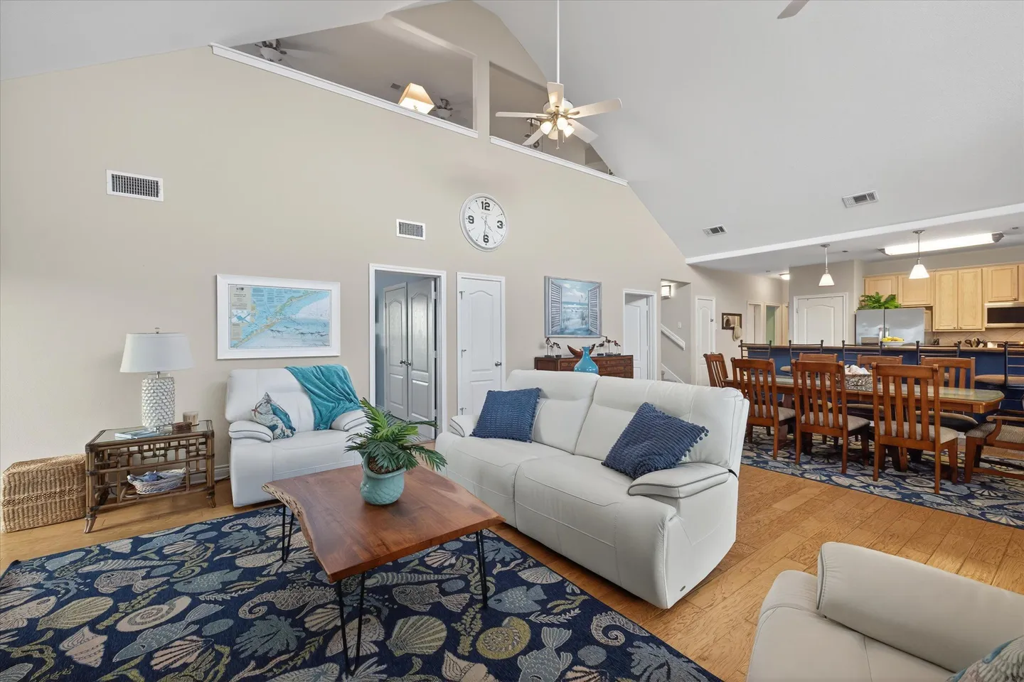Open-concept living room with vaulted ceilings, white sofas, patterned area rugs, and an adjacent dining area and kitchen.
