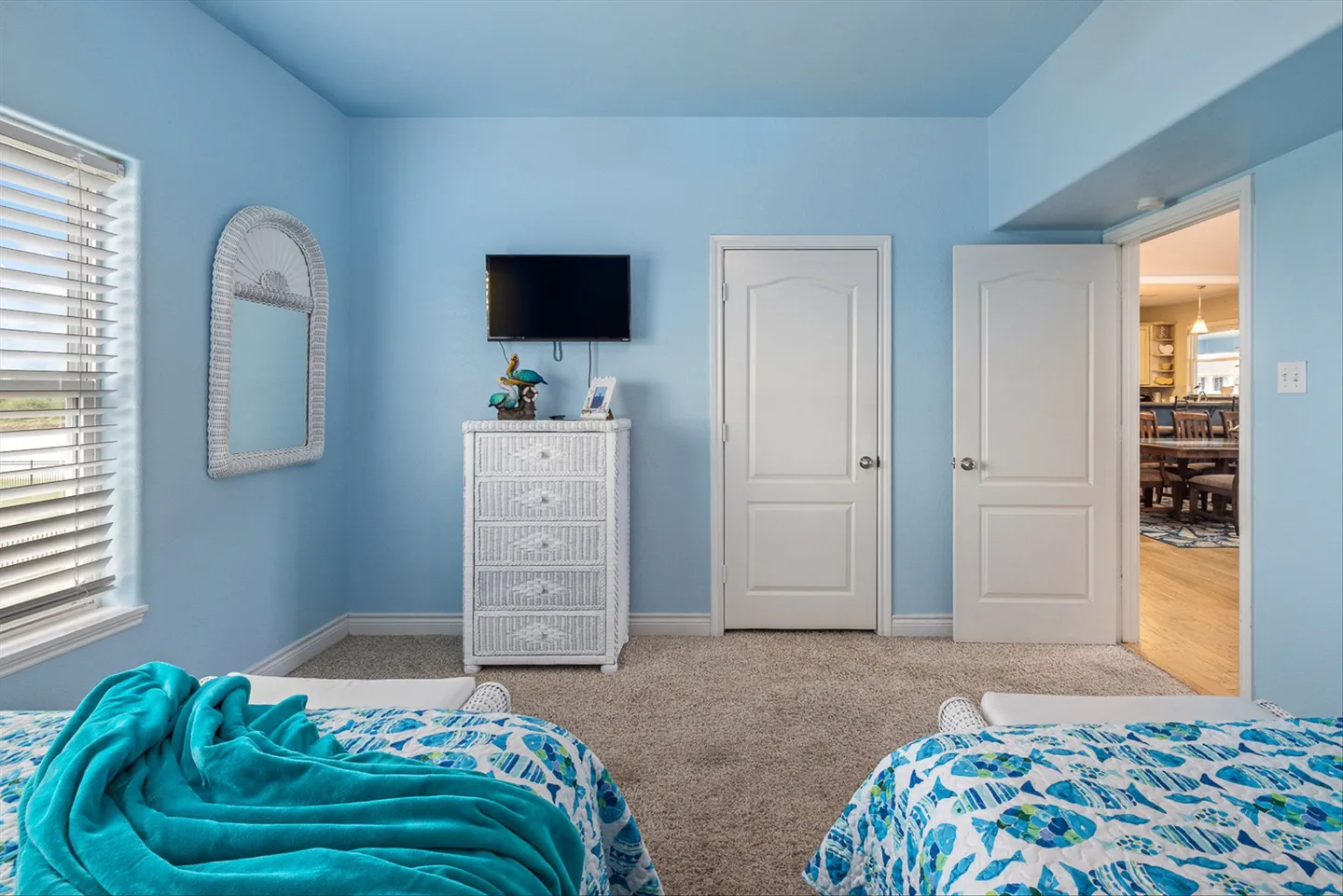 Bedroom with light blue walls, two twin beds with aquatic patterned bedding, a white wicker dresser, a wall-mounted TV, and carpeted flooring.