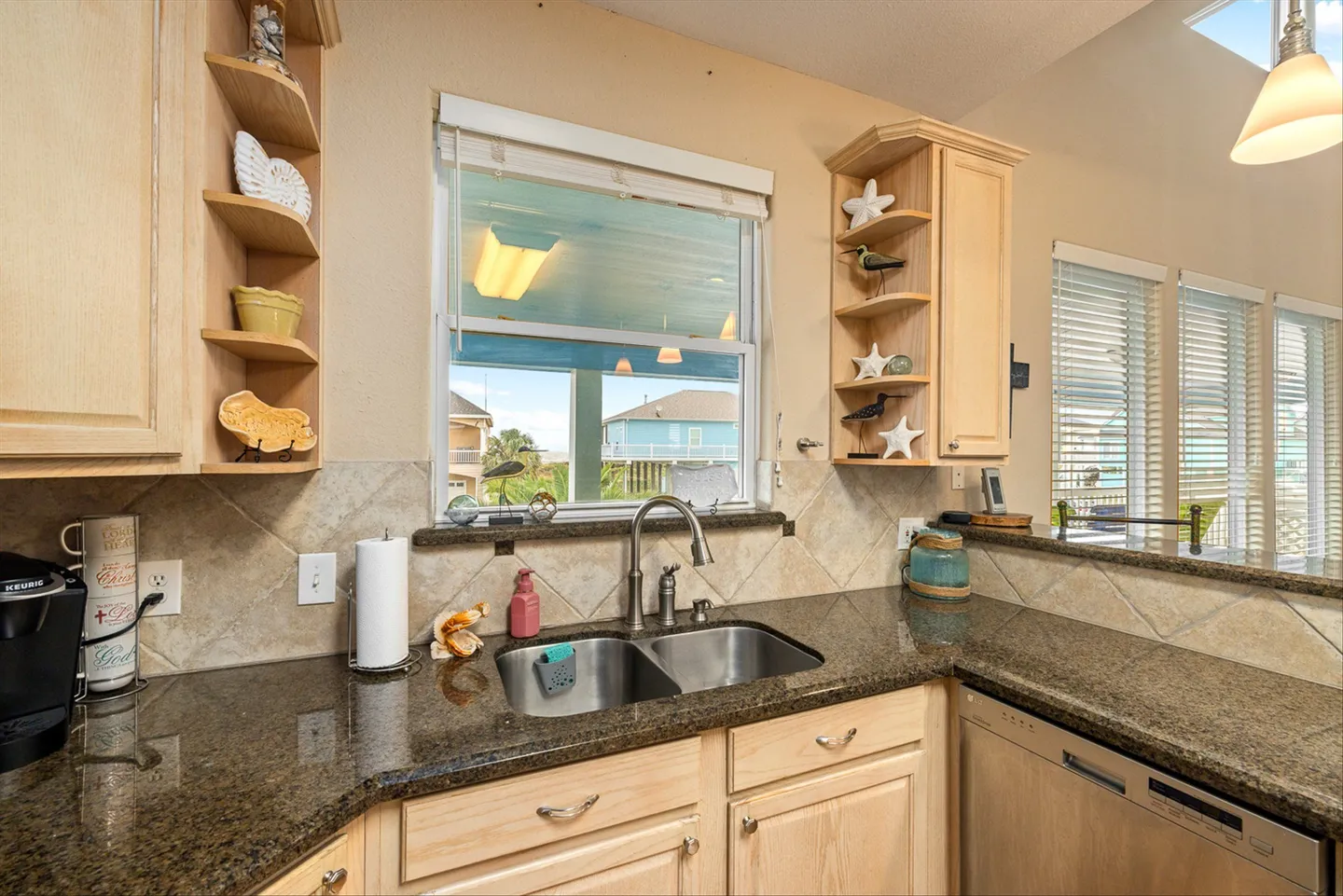 Kitchen counter and double sink beneath a window with views of nearby coastal houses, surrounded by light wood cabinetry and speckled countertops.