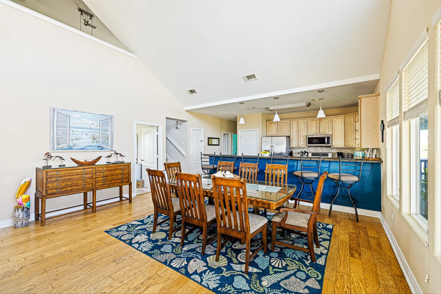 Dining area with a wooden dining table and chairs on a patterned rug, adjacent to a kitchen with a blue breakfast bar.